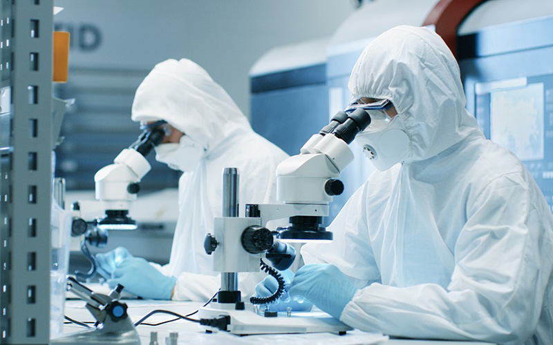 Kaksi insinööriä/tieteilijää/teknikkoa steriileissä puhdastilapukuissa käyttää mikroskooppeja komponenttien säätämiseen ja tutkimukseen.He työskentelevät elektroniikkakomponentteja valmistavassa tehtaassa.